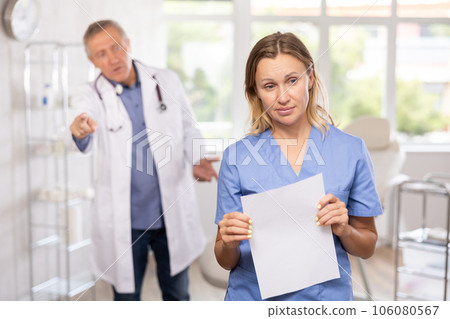 Frustrated female doctor with papers standing in medical office with angry senior colleague Frustrated female doctor with papers standing in medical office with angry senior colleague 106080567