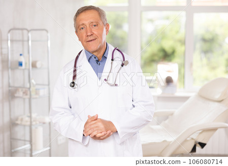 Old man doctor posing in doctor's cabinet with clinical chair 106080714