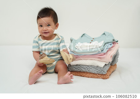 happy infant sitting with stack of baby clothes on bed 106082510