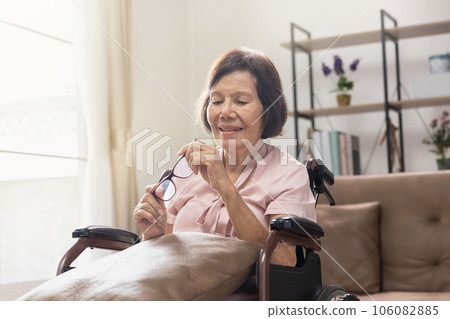Senior woman holding eyeglasses sitting on wheelchair at home Senior woman holding eyeglasses sitting on wheelchair at home 106082885