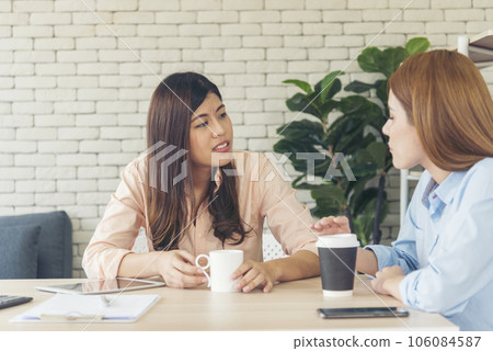 Two Women colleagues drinking black coffee cup at office desk together. Asian woman friendstalking smiling with friendship happy time. Teamwork happy moment lifestyle. International Woman day concept Two Women colleagues drinking black coffee cup at office desk together. Asian woman friendstalking smiling with friendship happy time. Teamwork happy moment lifestyle. International Woman day concept 106084587