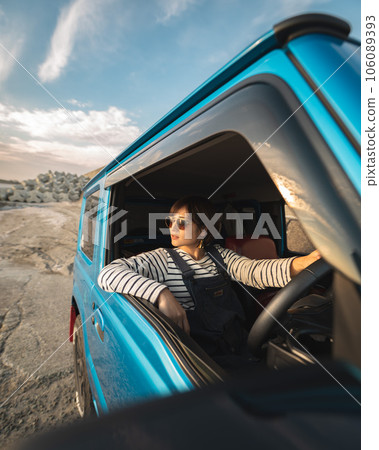 A woman in sunglasses sitting in the driver's seat along the seashore at sunset is looking into the distance with her elbows on the window 106089393