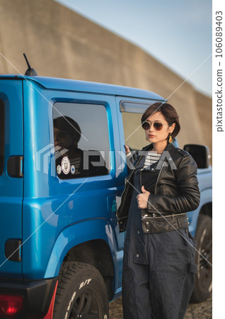 A woman in overalls and rider sunglasses is standing next to a car 106089403