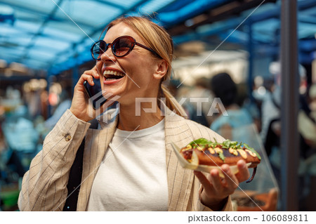 Smiling woman talking phone and demonstrate takeaway fast food hot dog while standing on food market 106089811