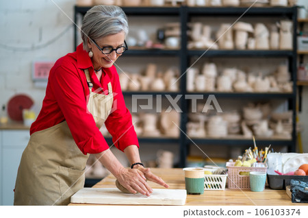 Mature woman wedging clay with hands standing behind table in studio. Mature woman wedging clay with hands standing behind table in studio. 106103374