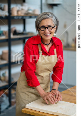 Mature woman wedging clay with hands standing behind table in studio. 106103376