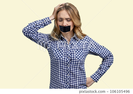 Portrait of young desperate blond woman with her mouth taped. Young lady touchine her head. Isolated on pastel yellow background. Portrait of young desperate blond woman with her mouth taped. Young lady touchine her head. Isolated on pastel yellow background. 106105496