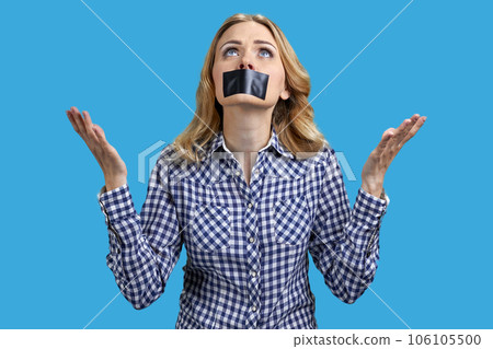 Portrait of a young woman with taped mouth is looking up. Attractive lady with her hands up. Isolated on vivid blue. Portrait of a young woman with taped mouth is looking up. Attractive lady with her hands up. Isolated on vivid blue. 106105500