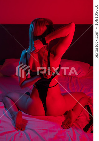 Back view of sexy blonde woman in leather bodysuit in bedroom in red blue neon light. Role-playing games. Back view of sexy blonde woman in leather bodysuit in bedroom in red blue neon light. Role-playing games. 106105610