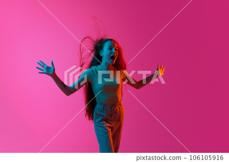 Young girl standing with wind blowing on hair, shouting, emotionally posing against pink studio background in neon light Young girl standing with wind blowing on hair, shouting, emotionally posing against pink studio background in neon light 106105916
