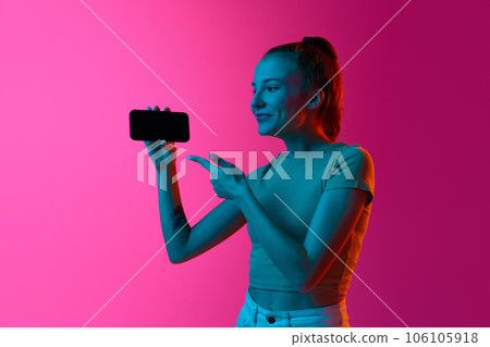 Smiling young girl pointing on mobile phone screen against pink studio background in neon light. Sales, online services Smiling young girl pointing on mobile phone screen against pink studio background in neon light. Sales, online services 106105918