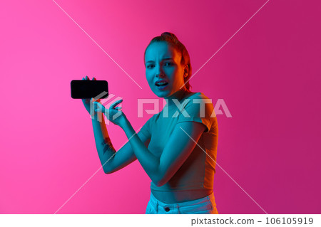 Young girl with doubtful, questioning face pointing on mobile phone screen against pink studio background in neon light. 106105919