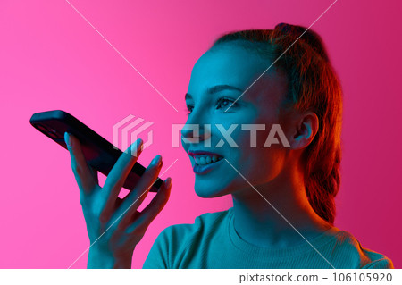 Cheerful, smiling girl recording voice message on mobile phone against pink studio background in neon light 106105920