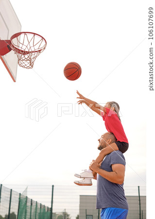 A child throws a ball into a basketball hoop, sitting on his father's shoulders. Dad teaches the child to play basketball. Outdoor sports ground A child throws a ball into a basketball hoop, sitting on his father's shoulders. Dad teaches the child to play basketball. Outdoor sports ground 106107699