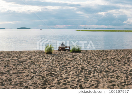 sandy beach on the shore of a vast lake, a woman traveler is sitting on a bench made of old pallets sandy beach on the shore of a vast lake, a woman traveler is sitting on a bench made of old pallets 106107937