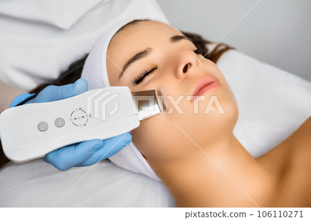 Expert in work uniform cleans woman client face with ultrasonic equipment. Portable appliance for improving quality of female skin concept 106110271