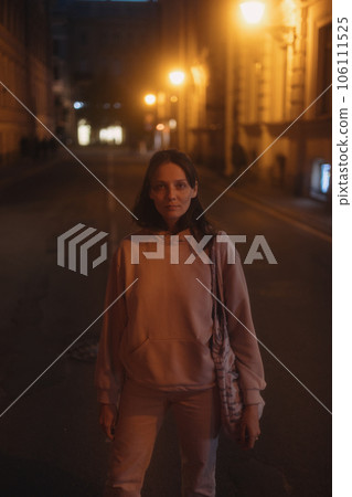 young thoughtful woman stands on road in beige hoodie. walk in evening in light of lanterns, 106111525