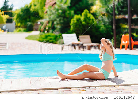 Cute and funny blonde teenage girl in a swimsuit sits on the side of the pool.  106112648