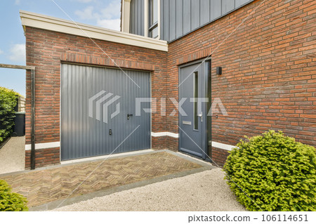 a grey garage door in front of a red brick wall with green bushes and shrubs on either side of the building 106114651