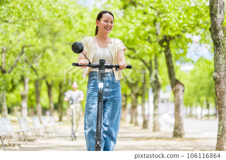 可愛的年輕女子帶著電動踢腳板在令人耳目一新的綠樹成蔭的街道上興奮地奔跑 | 電動踢腳板圖像 106116864