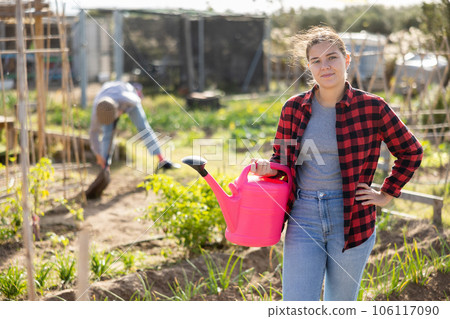 Skilled young woman farmer in plaid shirt holding pink watering can in hands and smiling at camera during work in vegetable garden in autumn 106117090