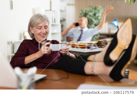 Relaxed elderly female office employee playing games on smartphone 106117579