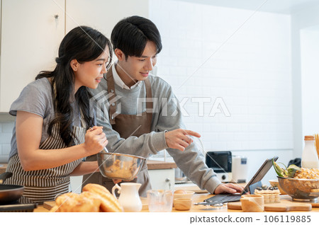 A lovely Asian couple is reading a pastry recipe on a tablet, enjoying cooking dates at home A lovely Asian couple is reading a pastry recipe on a tablet, enjoying cooking dates at home 106119885