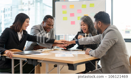 A multiracial group of cheerful business people is in a meeting, putting hands together as a team A multiracial group of cheerful business people is in a meeting, putting hands together as a team 106121469