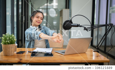 A happy Asian female radio host stretches her arms at her desk after finishing her live radio show A happy Asian female radio host stretches her arms at her desk after finishing her live radio show 106121889