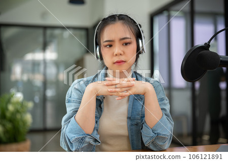 A stressed and serious Asian female radio host sits at her desk in the studio with an unhappy face 106121891