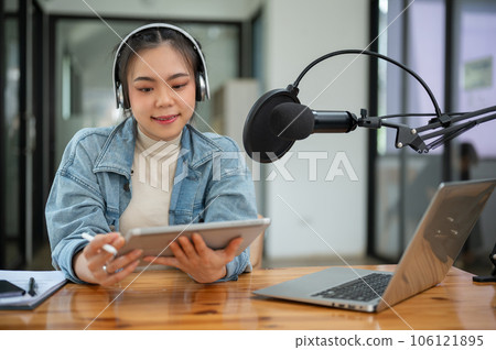 A beautiful Asian female radio host is looking at her tablet screen, speaking into a microphone A beautiful Asian female radio host is looking at her tablet screen, speaking into a microphone 106121895