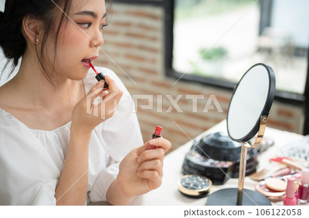 A beautiful Asian female applies lipstick to her lips, getting ready, put on makeup at home. 106122058