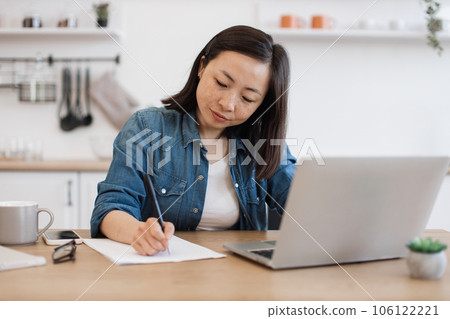 Woman making notes of article from computer in apartment Woman making notes of article from computer in apartment 106122221