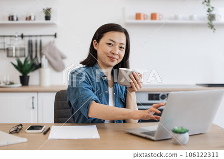 Adult posing with drink while working on laptop in kitchen Adult posing with drink while working on laptop in kitchen 106122311