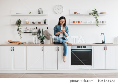 Lady using smartphone while sitting on countertop in kitchen 106123528