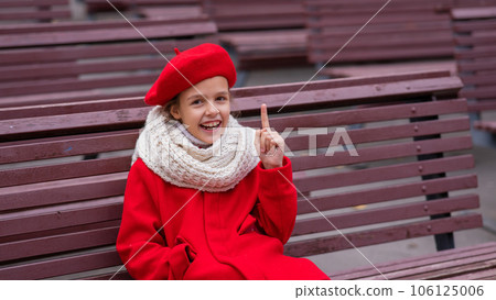 Smiling caucasian girl in a red coat and beret sits alone on a bench.  106125006
