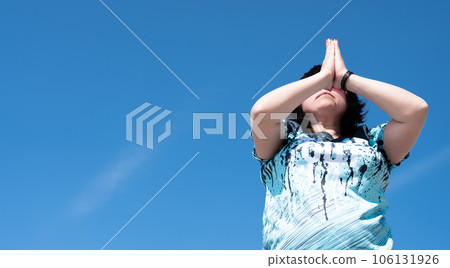 An impersonal portrait of a woman performing a set of yoga exercises, surya namaskara, holding her hands in front of her face against a blue sky 106131926