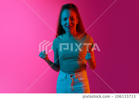 Young girl holding fists up, feeling worried and emotional against pink studio background in neon light 106133713