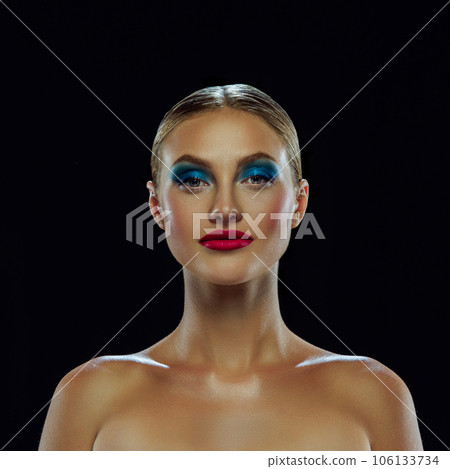 Portrait of young blond woman with bare shoulders and bright, unusual, creative make-up over dark mode background. Fashion model shoot. 106133734
