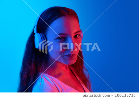 Smiling girl listening to music in headphones against blue studio background in neon light. Enjoyment and relaxation 106133735