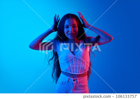 Young woman feeling cheerful and astonished, standing against blue studio background in neon light. Good and excited Young woman feeling cheerful and astonished, standing against blue studio background in neon light. Good and excited 106133757