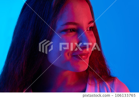Close-up of beautiful girl with little smile looking away against blue studio background in neon light. Good mood 106133765