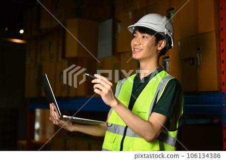 Male worker in hardhat and vest using laptop, checking inventory production stock control in warehouse 106134386