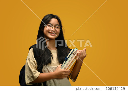 Smiling teenager schoolgirl with backpack holding books standing on yellow background. Back to school concept. Smiling teenager schoolgirl with backpack holding books standing on yellow background. Back to school concept. 106134429