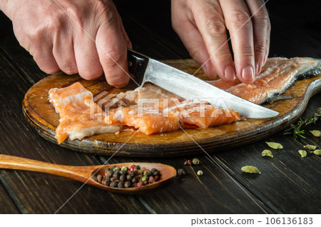 The hands of a cook cut raw salmon fish on a kitchen cutting board. Delicious red fish food for restaurant or hotel. The hands of a cook cut raw salmon fish on a kitchen cutting board. Delicious red fish food for restaurant or hotel. 106136183
