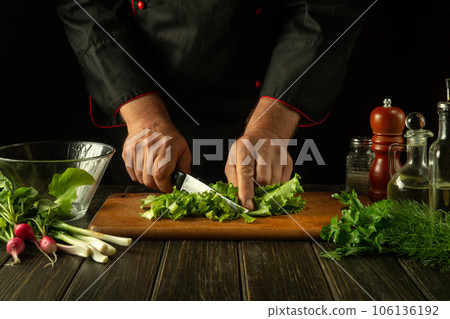 The cook prepares a salad in the restaurant kitchen. Close-up of a chef hands cutting Lettuce with a knife on a cutting board. Fresh vegetarian food on the kitchen table 106136192