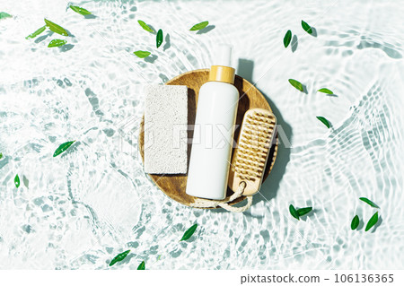 Body care essentials, daily routine background with white mockup tube, pumice stone and cosmetic brush in a wooden plate floating in clean water. 106136365