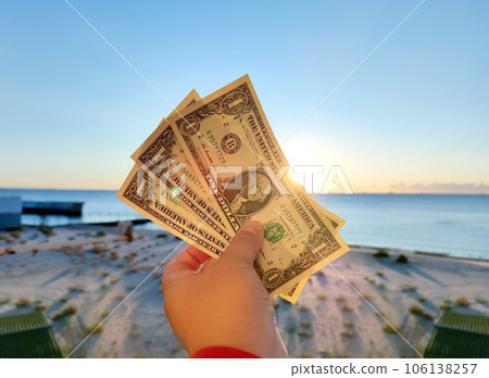 Person holding in hand three denominations of US dollars banknotes with face value of one dollar on background of sea waves on sandy beach of seashore on a sunny summer morning. Concept money finance Person holding in hand three denominations of US dollars banknotes with face value of one dollar on background of sea waves on sandy beach of seashore on a sunny summer morning. Concept money finance 106138257