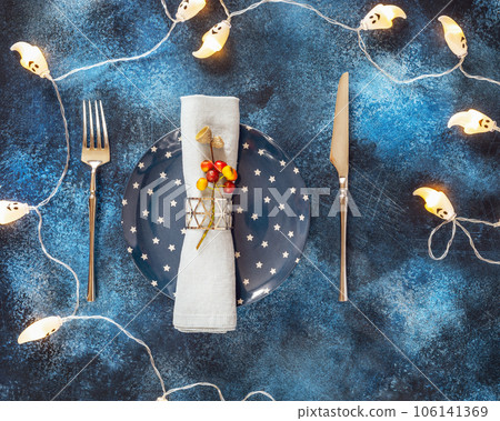 Halloween Dinner. Helloween holiday composition with festive plate with a knife, fork and napkin on a blue background with garland with ghosts. Table setting for Halloween Halloween Dinner. Helloween holiday composition with festive plate with a knife, fork and napkin on a blue background with garland with ghosts. Table setting for Halloween 106141369