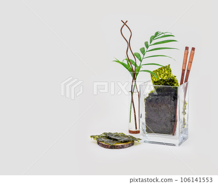 Organic dried seaweed snack in a glass jar with chopsticks and palm leaf on a blue background with copy space. Healthy sea snack suitable for keto and vegan diet. Japanese food, chips of nori seaweed 106141553